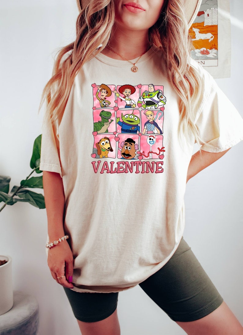 K&ouml;nnte beinhalten: Ein wei&szlig;es T-Shirt mit einem Aufdruck von Toy Story-Charakteren in rosafarbenen Herzen. Der Text "My Valentine" befindet sich unter den Figuren.