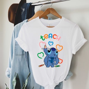 May include: White t-shirt featuring a blue Stitch character drawing hearts with colourful crayons. The text "teach" is written in rainbow colours above Stitch.