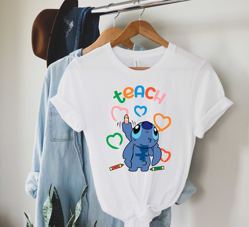 May include: White t-shirt featuring a blue Stitch character drawing hearts with colourful crayons. The text "teach" is written in rainbow colours above Stitch.