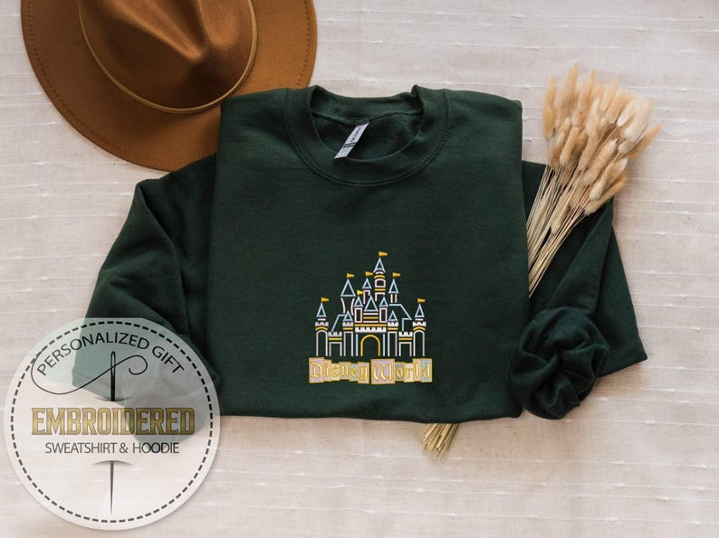 May include: Dark green sweatshirt with an embroidered castle design and the words "Disney World" in gold. The sweatshirt is folded on a light-colored surface, with a brown hat and dried flowers in the background.