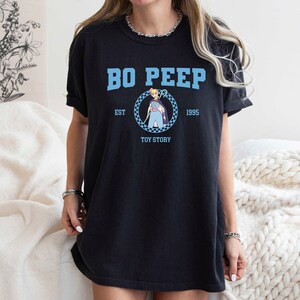 Puede incluir: Camiseta negra con un gr&aacute;fico azul de Bo Peep de Toy Story. El gr&aacute;fico incluye el texto "Bo Peep", "Est. 1995", y "Toy Story".