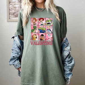 K&ouml;nnte beinhalten: Gr&uuml;nes T-Shirt mit einem Aufdruck der Toy Story-Charaktere in rosa Herzen. Der Text "VALENTINE" ist unter dem Aufdruck gedruckt.