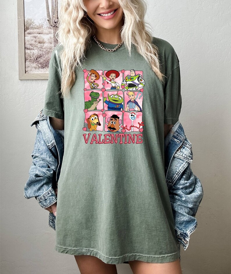 K&ouml;nnte beinhalten: Gr&uuml;nes T-Shirt mit einem Aufdruck der Toy Story-Charaktere in rosa Herzen. Der Text "VALENTINE" ist unter dem Aufdruck gedruckt.