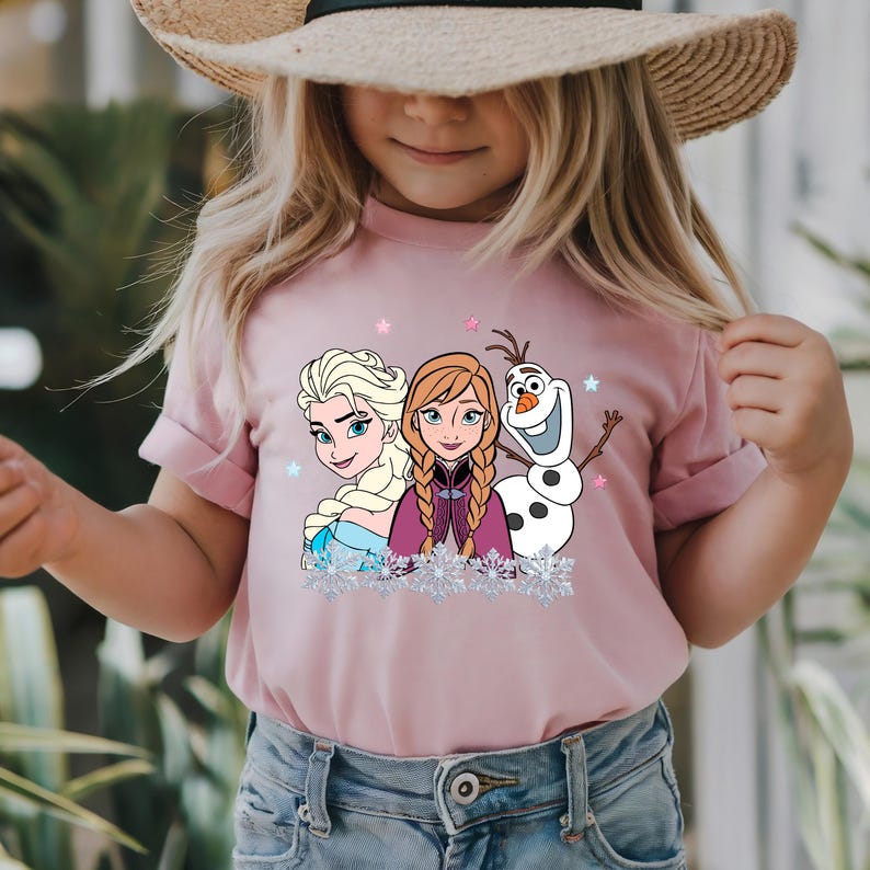 May include: A light pink t-shirt featuring Elsa, Anna, and Olaf from the movie Frozen. The graphic includes snowflakes and stars. The shirt is short-sleeved and appears to be made of a soft material.