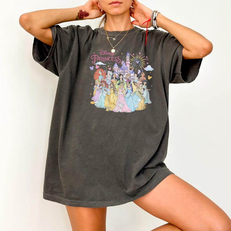 May include: Dark gray t-shirt featuring a colorful graphic of Disney Princesses with a castle and fireworks. The text "Disney Princess" is above the graphic. The shirt has short sleeves and a relaxed fit.