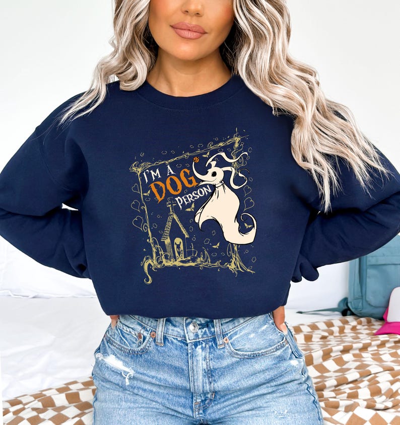 May include: Navy blue sweatshirt with a graphic featuring the text "I'M A DOG PERSON" and a cartoon character. The design includes a house, bats, and a ghost-like figure in a decorative frame. The sweatshirt is a casual style.