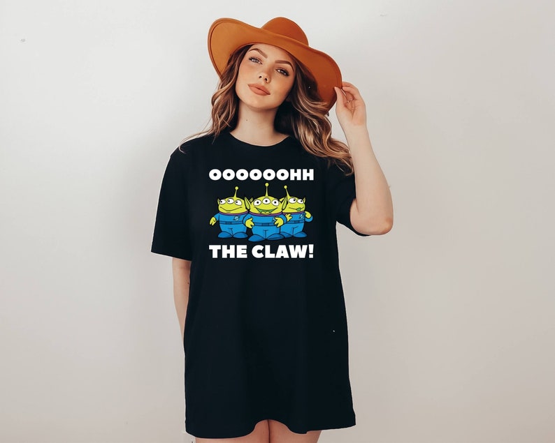 K&ouml;nnte beinhalten: Schwarzes T-Shirt mit einem Aufdruck von drei gr&uuml;nen Au&szlig;erirdischen mit dem Text "Oooooooooh The Claw!" auf der Vorderseite.