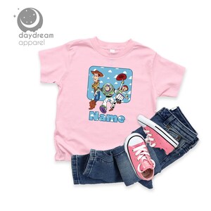 May include: Pink t-shirt with a cartoon image of Woody, Buzz Lightyear, and Jessie from Toy Story. The shirt has a blue square with white clouds and the text "Name" below the image.