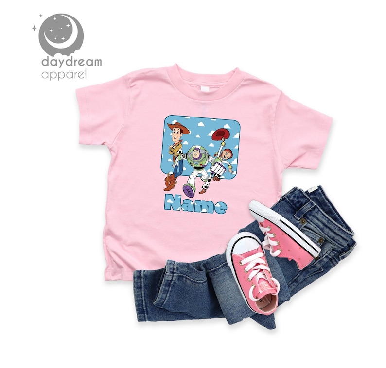 May include: Pink t-shirt with a cartoon image of Woody, Buzz Lightyear, and Jessie from Toy Story. The shirt has a blue square with white clouds and the text "Name" below the image.