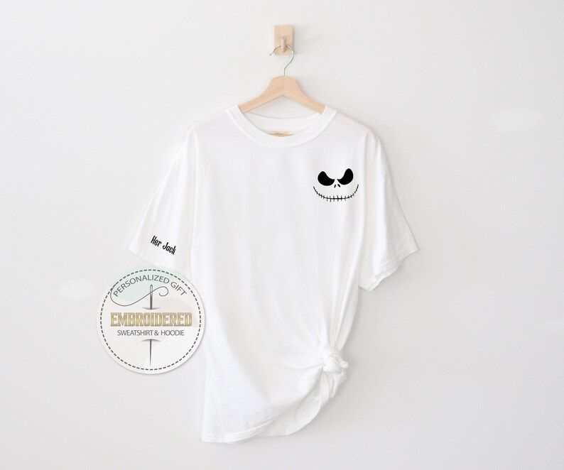 K&ouml;nnte beinhalten: Wei&szlig;es T-Shirt mit einem schwarzen Jack Skellington-Gesicht auf der Brust. Auf dem linken &Auml;rmel steht der gestickte Text "Her Jack". Das Shirt h&auml;ngt an einem Holzb&uuml;gel vor wei&szlig;em Hintergrund. Ein kreisf&ouml;rmiges Logo ist sichtbar.