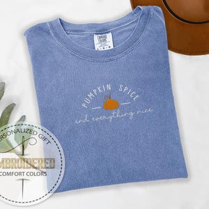 May include: A blue Comfort Colors t-shirt with the embroidered phrase "Pumpkin Spice and everything nice" and a small orange pumpkin graphic. A brown hat is visible in the background. The shirt is a personalized gift.