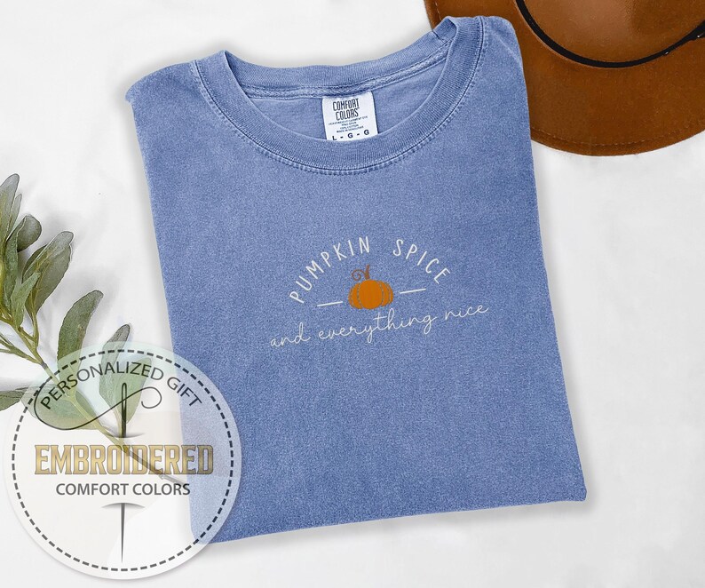 May include: A blue Comfort Colors t-shirt with the embroidered phrase "Pumpkin Spice and everything nice" and a small orange pumpkin graphic. A brown hat is visible in the background. The shirt is a personalized gift.