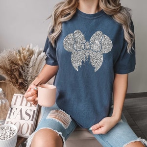 Puede incluir: Una camiseta azul marino con un lazo de purpurina plateado y un dise&ntilde;o de castillo. La camiseta es de gran tama&ntilde;o y tiene mangas cortas.