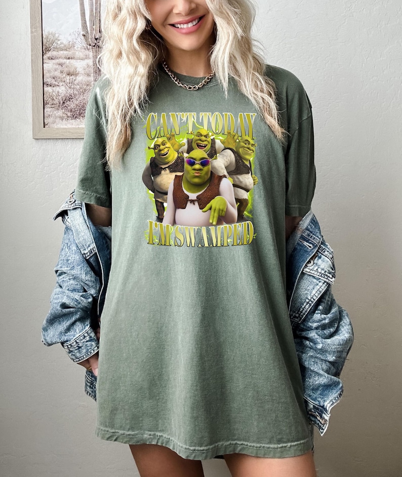 May include: Olive green t-shirt with a Shrek graphic and the text "CAN'T TODAY I'M SWAMPED". The graphic shows Shrek wearing sunglasses and other characters. The shirt has short sleeves and a relaxed fit.