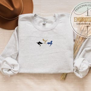 May include: A light gray sweatshirt with embroidered dragon silhouettes in black, gold, and blue. The text "PERSONALIZED GIFT EMBROIDERED SWEATSHIRT & HOODIE" is in a circular design. A brown hat and dried grass are also in the image.