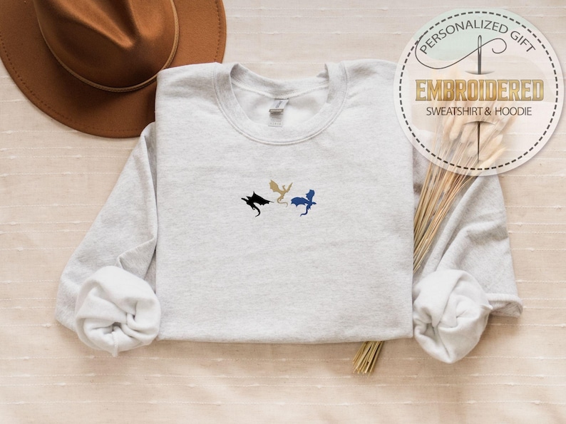 May include: A light gray sweatshirt with embroidered dragon silhouettes in black, gold, and blue. The text "PERSONALIZED GIFT EMBROIDERED SWEATSHIRT & HOODIE" is in a circular design. A brown hat and dried grass are also in the image.