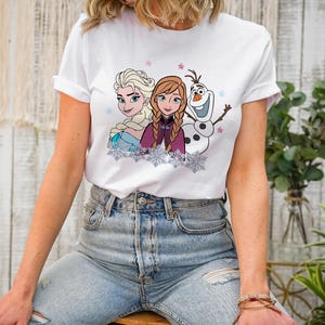 May include: White t-shirt featuring a colorful graphic of Elsa, Anna, and Olaf from the movie Frozen. The design includes snowflakes and stars. The shirt is made of a soft material.