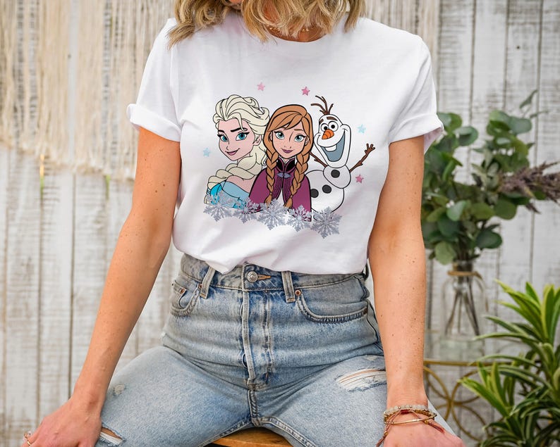 May include: White t-shirt featuring a colorful graphic of Elsa, Anna, and Olaf from the movie Frozen. The design includes snowflakes and stars. The shirt is made of a soft material.