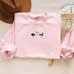 May include: A light pink sweatshirt with three embroidered dragon silhouettes in black, gold, and blue. The sweatshirt has long sleeves and a crew neck. The image also includes a brown hat and text that says "PERSONALIZED GIFT EMBROIDERED SWEATSHIRT & HOODIE".