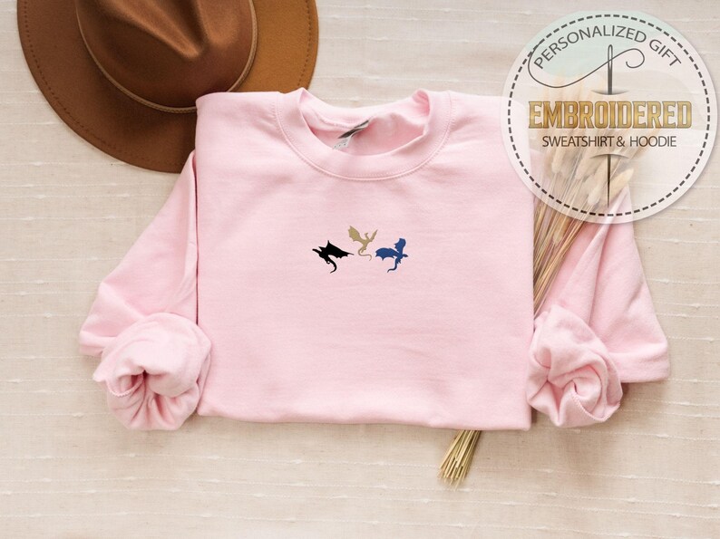 May include: A light pink sweatshirt with three embroidered dragon silhouettes in black, gold, and blue. The sweatshirt has long sleeves and a crew neck. The image also includes a brown hat and text that says "PERSONALIZED GIFT EMBROIDERED SWEATSHIRT & HOODIE".