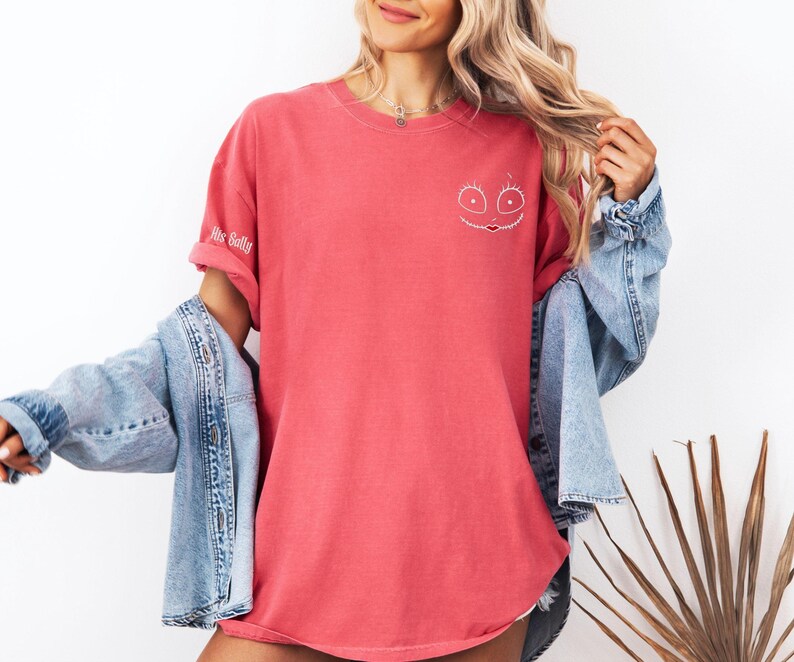 K&ouml;nnte beinhalten: Korallfarbenes T-Shirt mit wei&szlig;em, gesticktem Gesichtsdesign und dem Text "His Sally" auf dem &Auml;rmel. Das Shirt wird mit einer hellblauen Jeansjacke getragen.