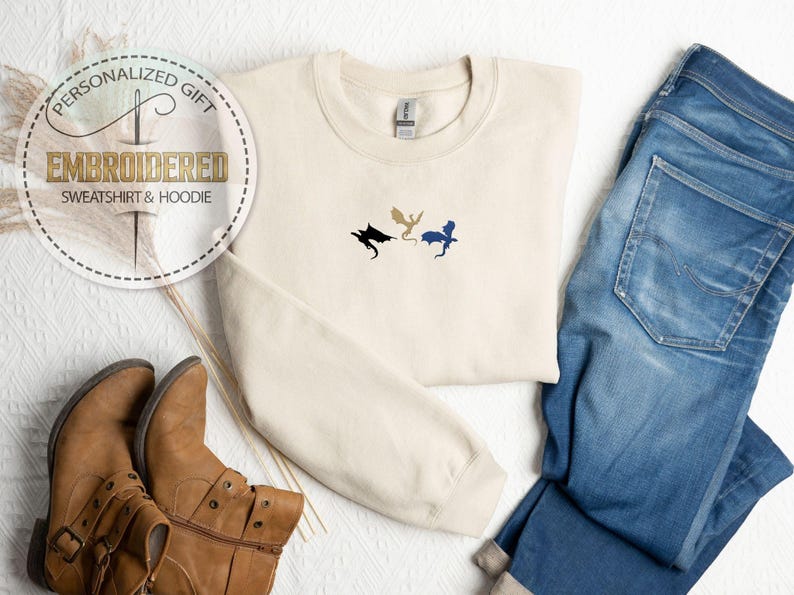 May include: Cream-colored embroidered sweatshirt with three dragon silhouettes in black, gold, and blue. The sweatshirt is paired with blue jeans and brown boots. The image also includes a circular graphic with text that reads "Personalized Gift EMBROIDERED SWEATSHIRT & HOODIE."