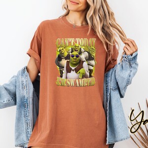 May include: A terracotta-coloured t-shirt with a graphic of Shrek and his friends, and the text "CAN'T TODAY I'M SWAMPIN'". The design includes Shrek wearing sunglasses. The shirt has rolled-up sleeves and a relaxed fit.