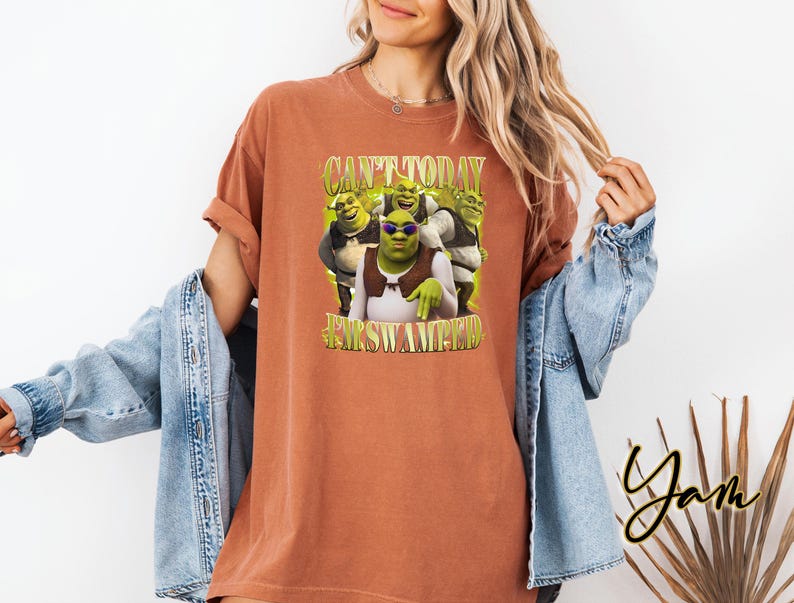 May include: A terracotta-coloured t-shirt with a graphic of Shrek and his friends, and the text "CAN'T TODAY I'M SWAMPIN'". The design includes Shrek wearing sunglasses. The shirt has rolled-up sleeves and a relaxed fit.