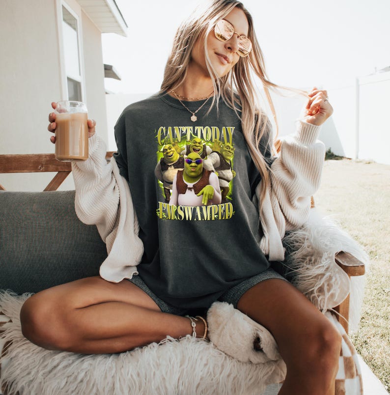 May include: Dark grey t-shirt with a Shrek graphic and the text "Can't Today, I'm Swamped" in yellow and green. The image shows Shrek wearing sunglasses with other characters from the movie. The shirt is worn by a person.