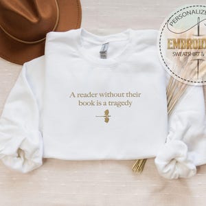 May include: White sweatshirt with the embroidered quote, "A reader without their book is a tragedy." The text is in a gold color, along with a small graphic. A brown hat is visible in the background. The sweatshirt is a personalized gift.