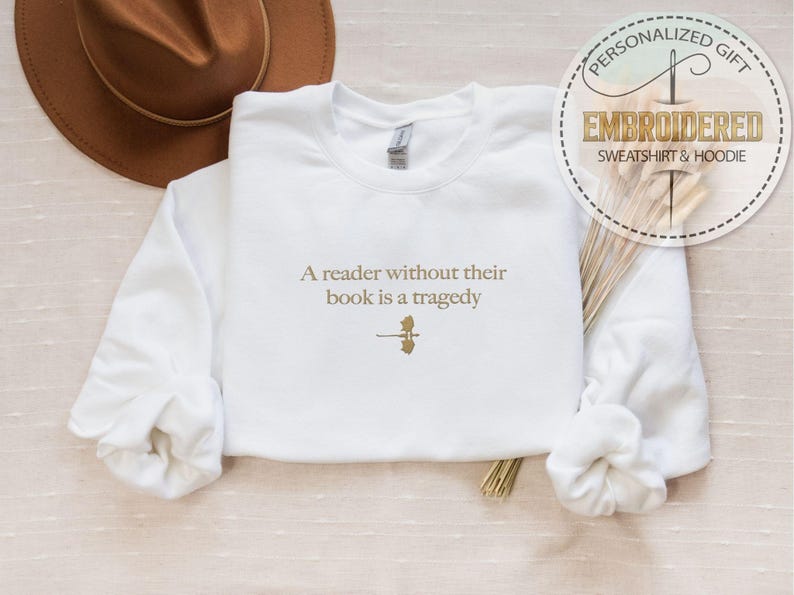 May include: White sweatshirt with the embroidered quote, "A reader without their book is a tragedy." The text is in a gold color, along with a small graphic. A brown hat is visible in the background. The sweatshirt is a personalized gift.