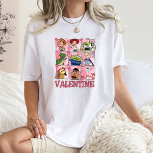 K&ouml;nnte beinhalten: Wei&szlig;es T-Shirt mit einem rosa und wei&szlig;en Aufdruck von Toy Story-Charakteren in einem Raster mit Herzen und dem Text "VALENTINE".