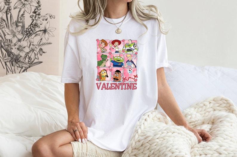 K&ouml;nnte beinhalten: Wei&szlig;es T-Shirt mit einem rosa und wei&szlig;en Aufdruck von Toy Story-Charakteren in einem Raster mit Herzen und dem Text "VALENTINE".