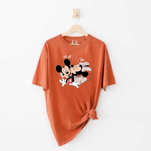 May include: A rust-coloured t-shirt featuring a graphic of Mickey Mouse and Minnie Mouse kissing. The graphic is surrounded by small pink hearts.