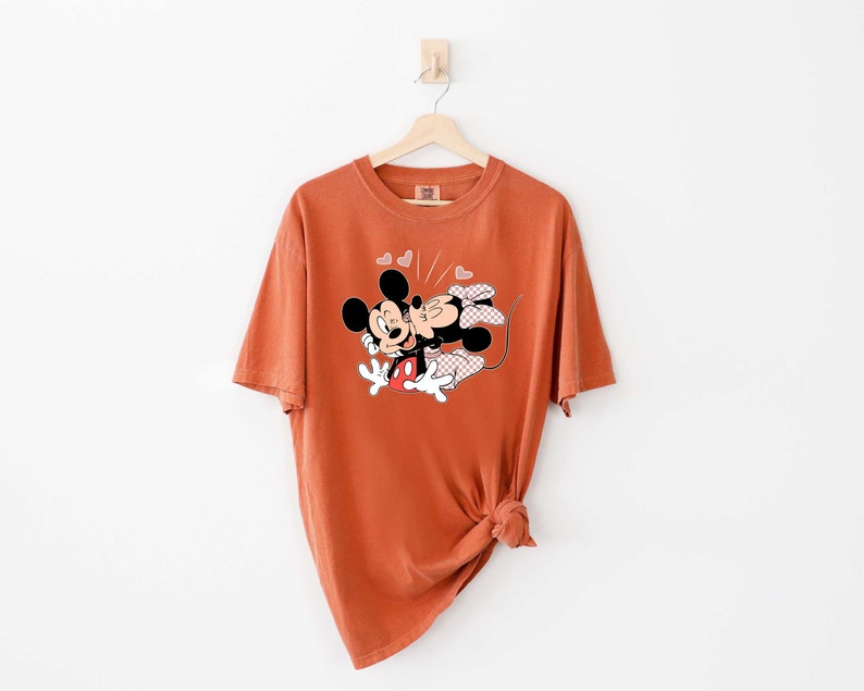 May include: A rust-coloured t-shirt featuring a graphic of Mickey Mouse and Minnie Mouse kissing. The graphic is surrounded by small pink hearts.