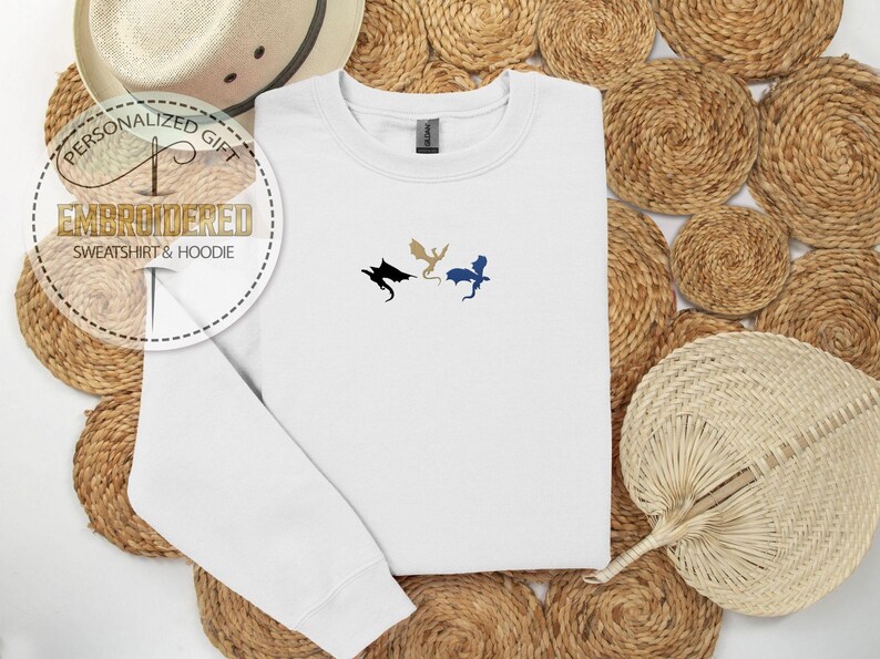 May include: White embroidered sweatshirt with three dragon silhouettes in black, gold, and blue. The sweatshirt is a crew neck style. The text "PERSONALIZED GIFT EMBROIDERED SWEATSHIRT & HOODIE" is in a circular design.
