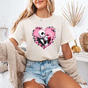 May include: Cream-colored t-shirt featuring a heart-shaped design with Jack Skellington and Sally from The Nightmare Before Christmas. The heart is surrounded by pink roses and the text "Love You to Death".