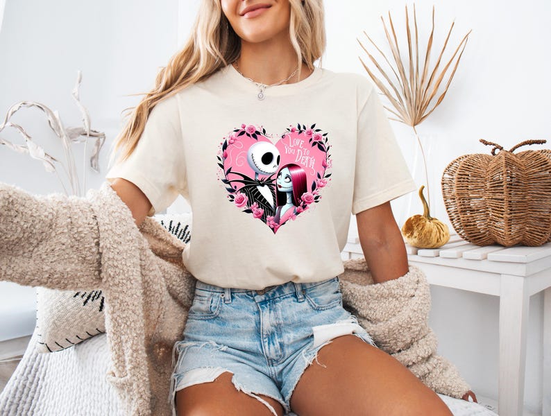 May include: Cream-colored t-shirt featuring a heart-shaped design with Jack Skellington and Sally from The Nightmare Before Christmas. The heart is surrounded by pink roses and the text "Love You to Death".