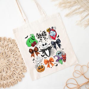 May include: A cream-colored tote bag featuring colorful illustrations of characters and bows from a popular animated film. The design includes Jack Skellington, Sally, and Oogie Boogie, with bats and spiderwebs. The bag has two handles.
