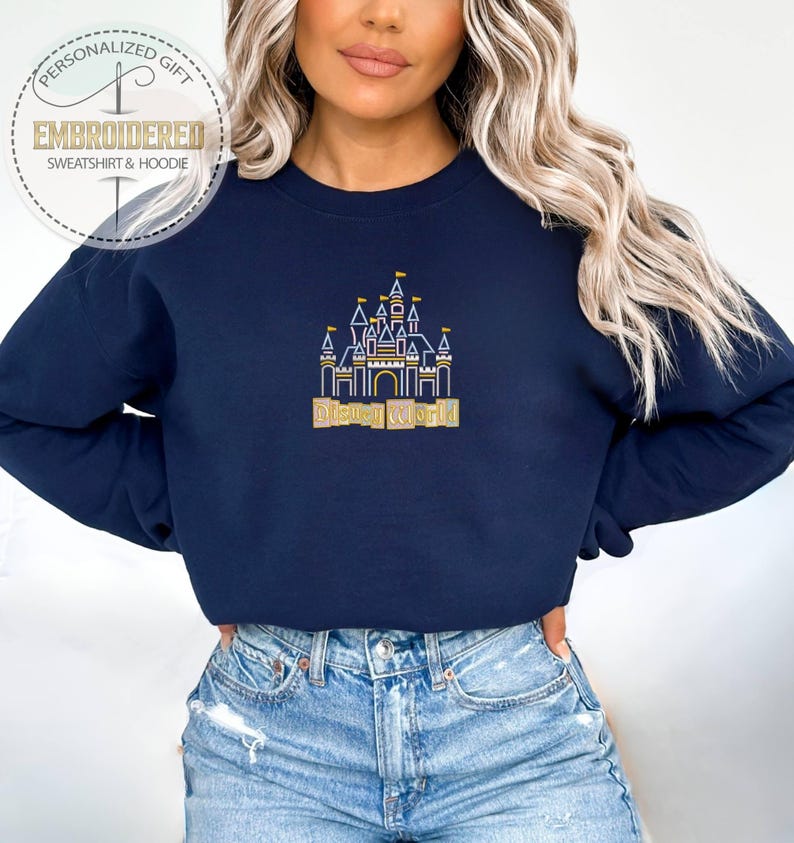 May include: Navy blue sweatshirt with an embroidered gold and white castle design and the words "Disney World". The sweatshirt has a crew neck and long sleeves. The image also includes a logo that says "Personalized Gift Embroidered Sweatshirt & Hoodie".