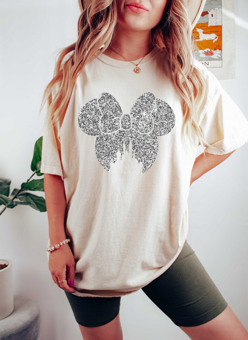 Puede incluir: Una camiseta blanca de gran tama&ntilde;o con un dise&ntilde;o de lazo de purpurina plateada. La camiseta la lleva puesta una persona con el pelo largo y rubio.