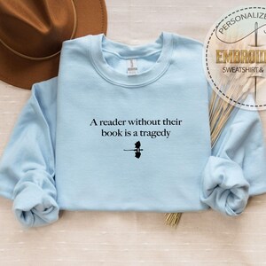 May include: A light blue sweatshirt with the quote "A reader without their book is a tragedy" in black text. The sweatshirt has long sleeves and a crew neck. A brown hat is visible in the upper left corner.