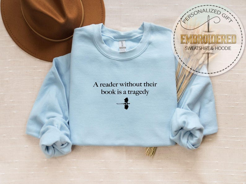May include: A light blue sweatshirt with the quote "A reader without their book is a tragedy" in black text. The sweatshirt has long sleeves and a crew neck. A brown hat is visible in the upper left corner.