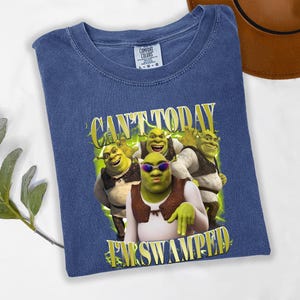 May include: A blue t-shirt with a graphic of the Shrek characters and the text "CAN'T TODAY I'M SWAMPED" in yellow. The shirt is made of a soft material and has a crew neck.