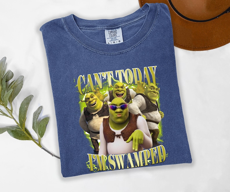 May include: A blue t-shirt with a graphic of the Shrek characters and the text "CAN'T TODAY I'M SWAMPED" in yellow. The shirt is made of a soft material and has a crew neck.
