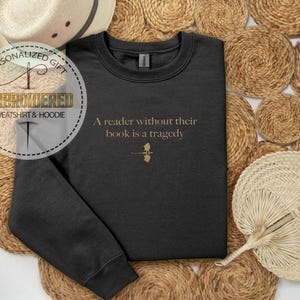 May include: Black sweatshirt with the quote "A reader without their book is a tragedy" in gold lettering. The sweatshirt has a small gold graphic below the text. The image also includes a straw hat and woven placemats.