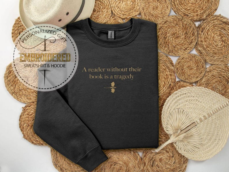May include: Black sweatshirt with the quote "A reader without their book is a tragedy" in gold lettering. The sweatshirt has a small gold graphic below the text. The image also includes a straw hat and woven placemats.