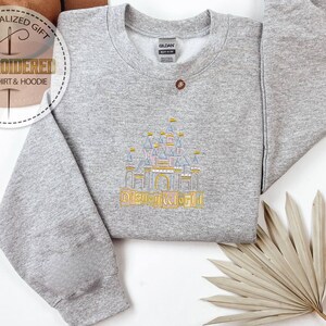 May include: A gray sweatshirt with an embroidered design of a castle and the text "DISNEYWORLD" in gold. The sweatshirt has a crew neck and long sleeves. The image also includes the text "PERSONALIZED GIFT" and "EMBROIDERED SWEATSHIRT & HOODIE".