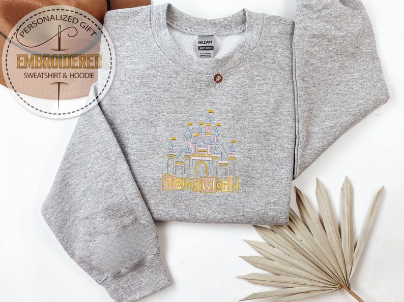 May include: A gray sweatshirt with an embroidered design of a castle and the text "DISNEYWORLD" in gold. The sweatshirt has a crew neck and long sleeves. The image also includes the text "PERSONALIZED GIFT" and "EMBROIDERED SWEATSHIRT & HOODIE".