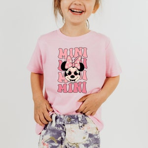 K&ouml;nnte beinhalten: Ein pinkfarbenes T-Shirt mit einem Aufdruck von Minnie Mouse, die eine Sonnenbrille und eine Schleife mit Blumen tr&auml;gt. Der Text "MINI" wird viermal auf dem Shirt wiederholt.